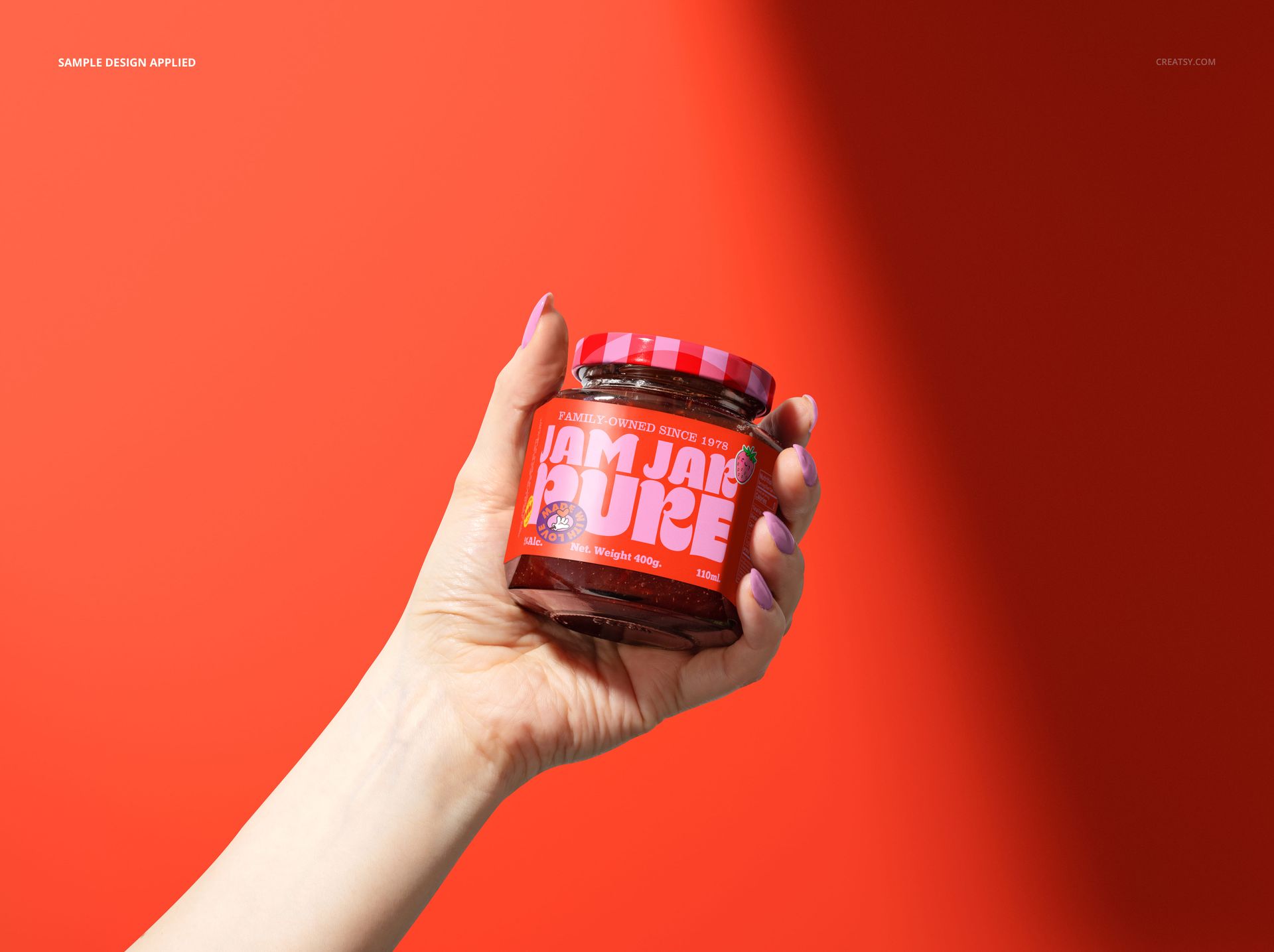 Glass jam jar with red label and checkered lid held in a hand against a solid bright red background, front view.