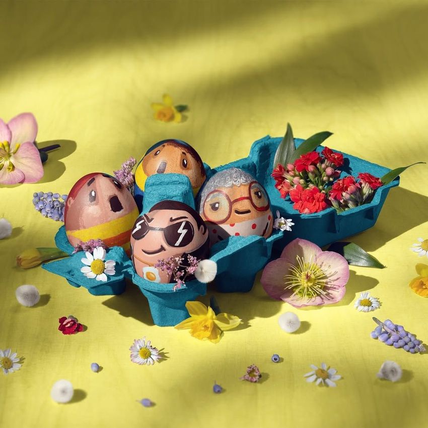 Egg carton mockup with four decorated eggs painted as cartoon faces, surrounded by colorful spring flowers on a yellow surface.