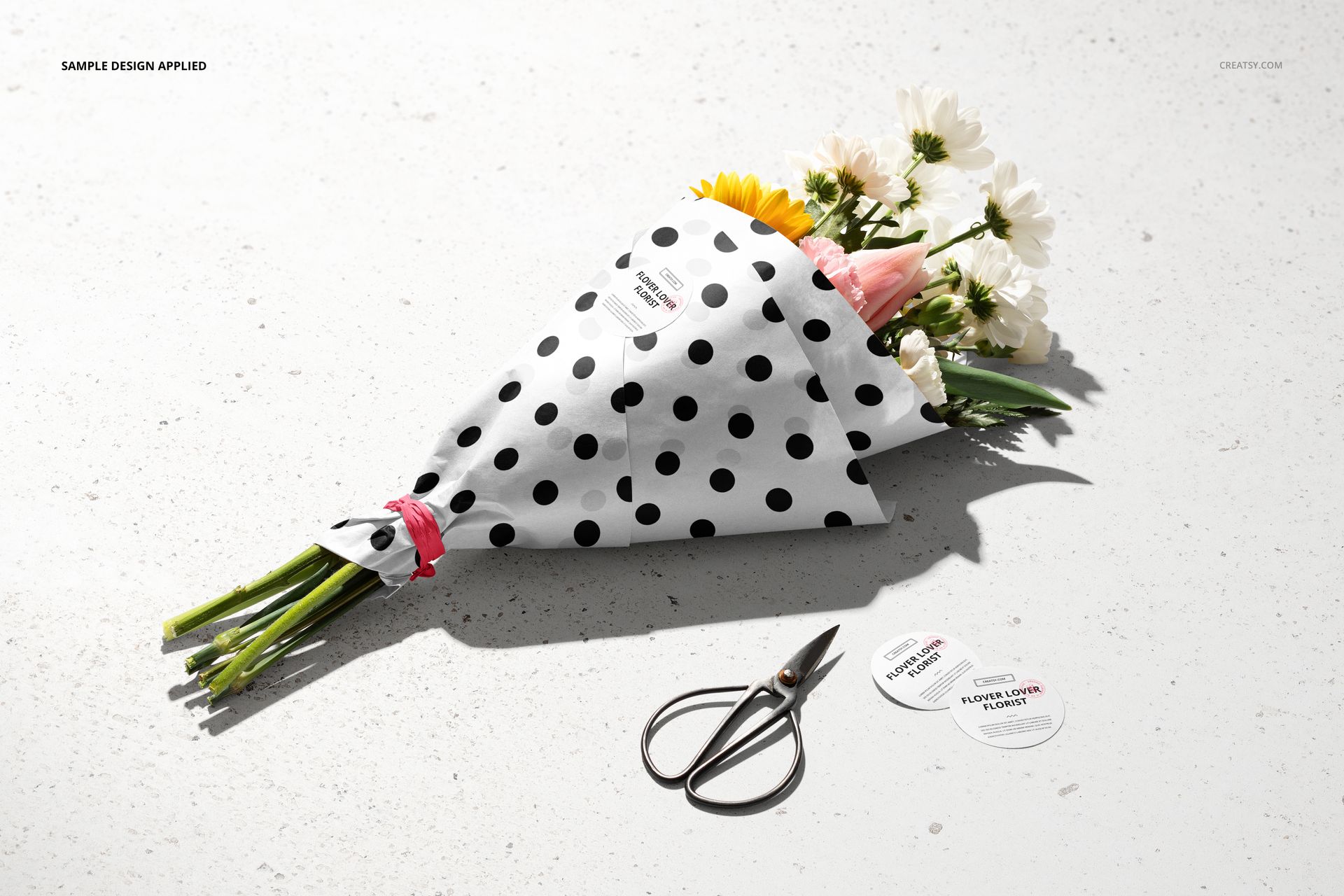 Flowers packaging mockup featuring a wrapped bouquet with black and white dotted paper, round stickers, and scissors on a textured surface.