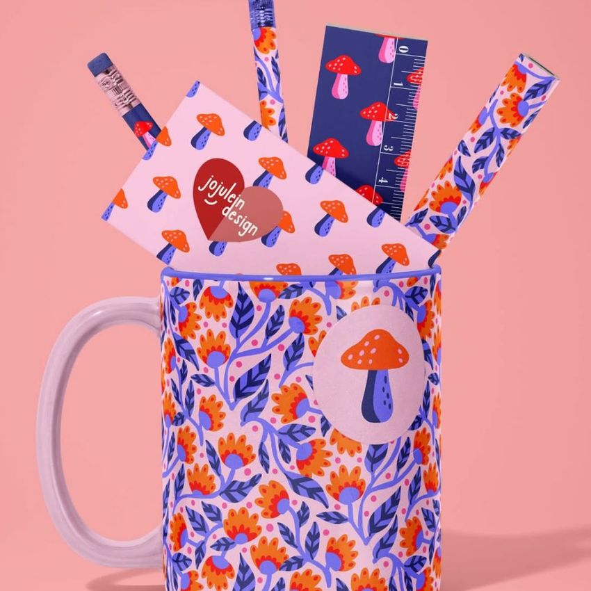 Mug mockup featuring a floral pattern, filled with office supplies like pencils, a ruler, and cards with mushroom designs on a pink background.