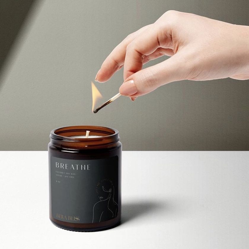 Brown glass candle jar with a black sticker mockup label, being lit by a hand holding a match, on a plain surface.