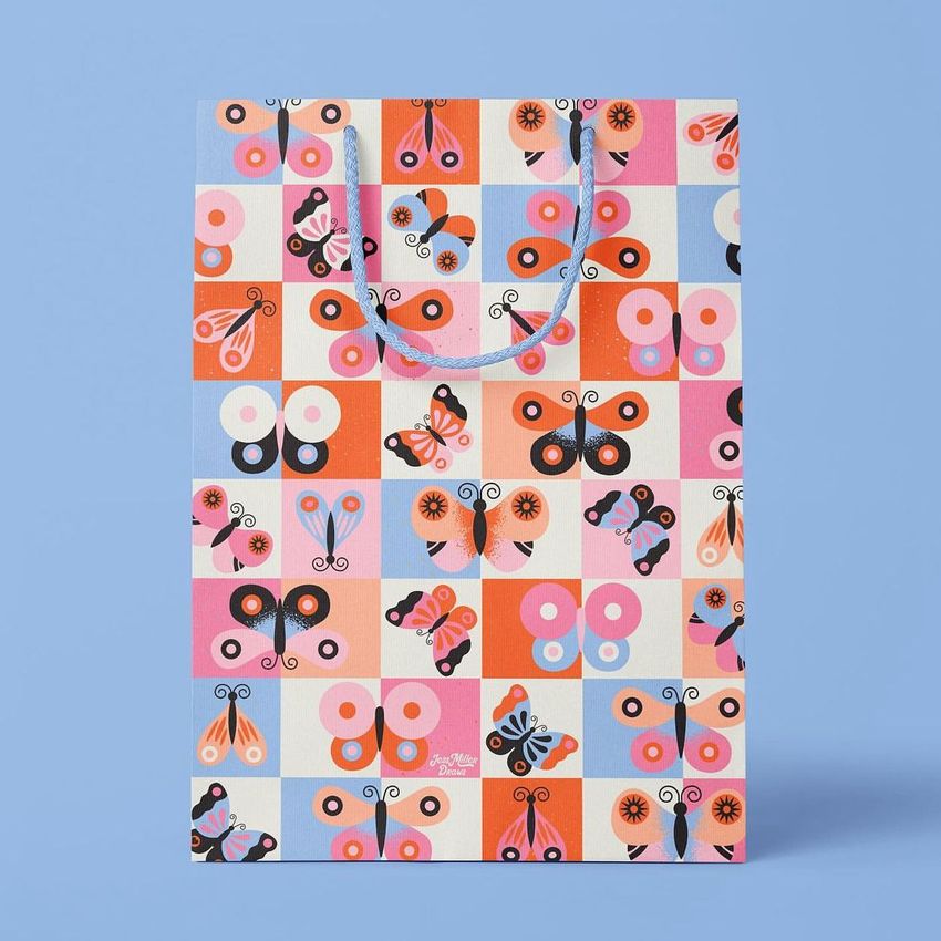 Paper bag mockup featuring a colorful butterfly pattern in a grid layout, displayed upright on a solid light blue background.