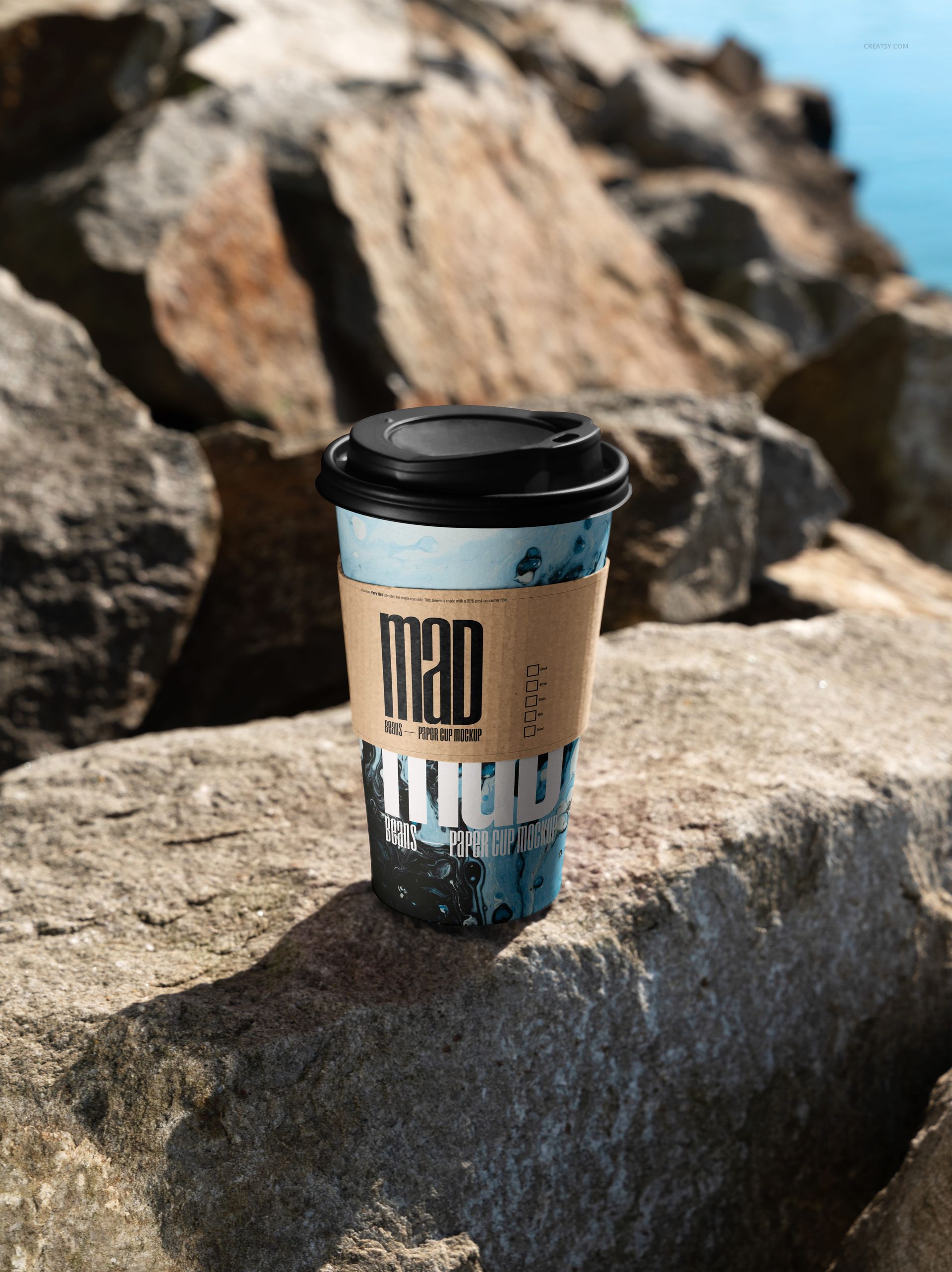PSD mockup of a paper coffee cup featuring a black lid and kraft sleeve, positioned on rugged shoreline rocks.