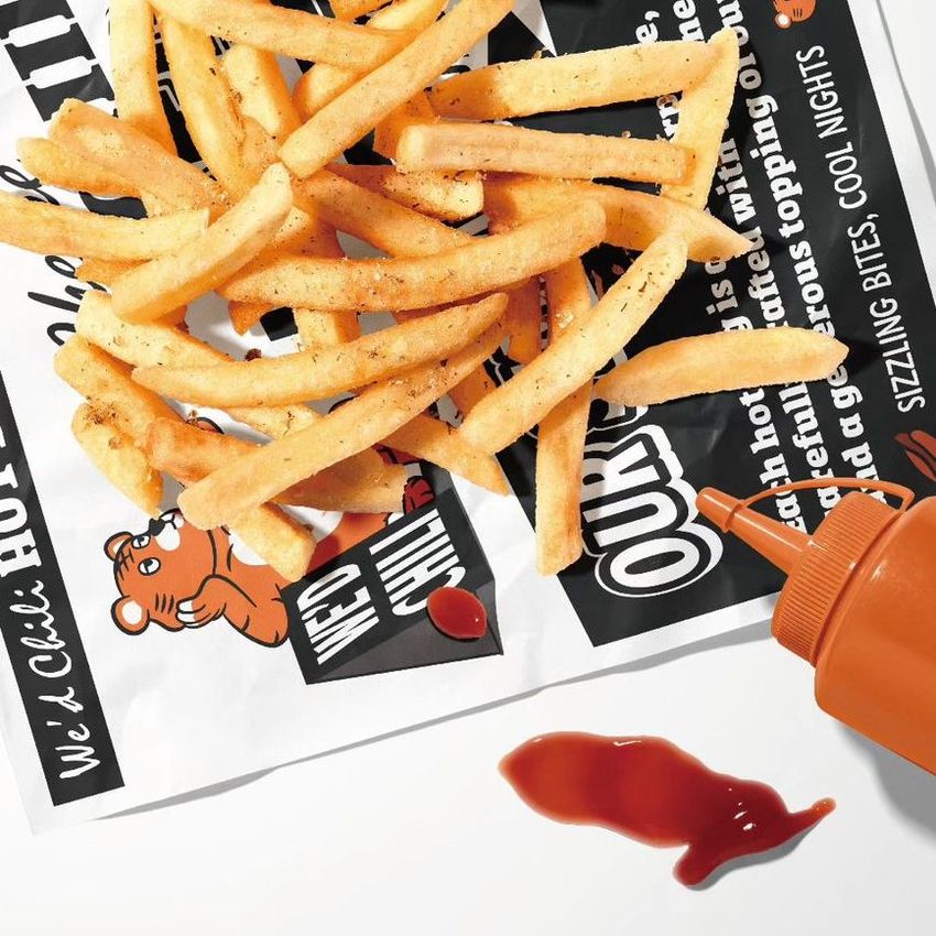 French fries are scattered on printed food safe paper mockup with bold black and orange text, next to an orange sauce bottle and ketchup spill.