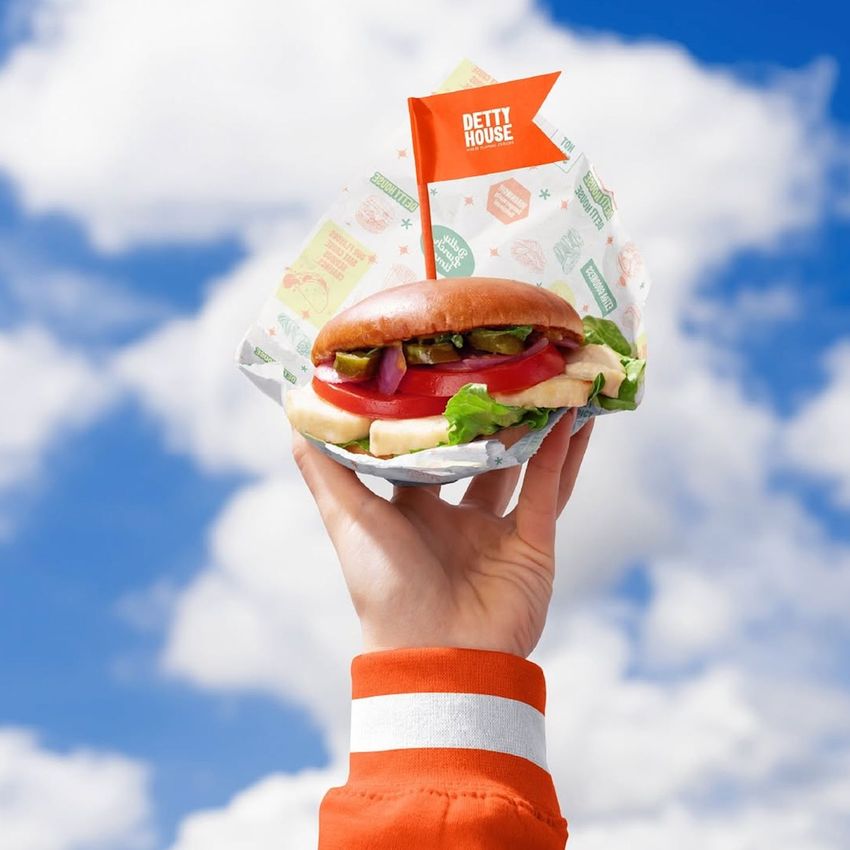 Vegan burger bar packaging mockup showing a hand holding a sandwich with printed wrap and a visible orange flag label.