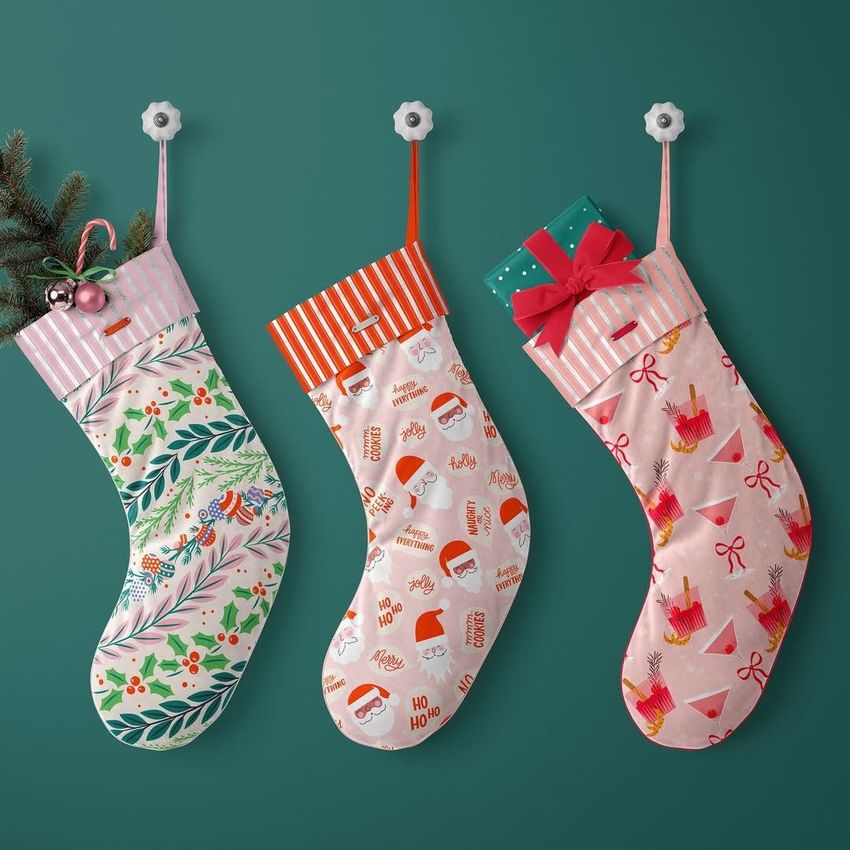 Three velvet Christmas stocking mockups hang on a green wall, each with different festive patterns and filled with holiday items.