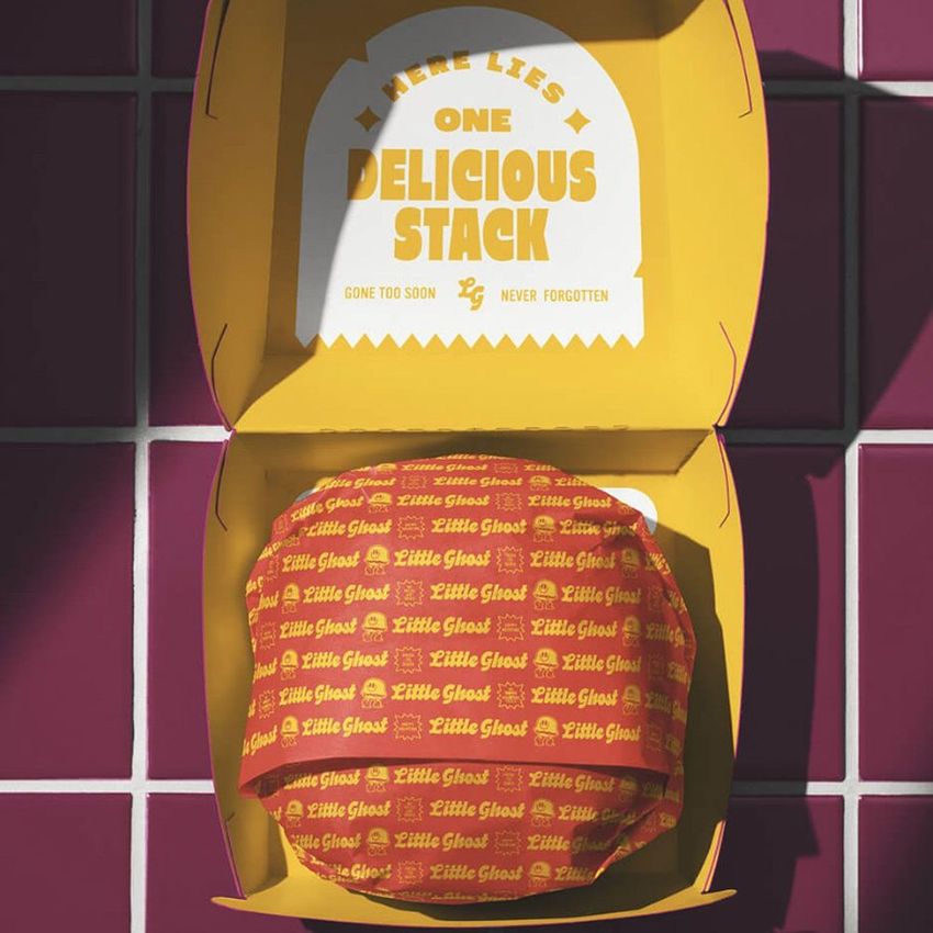 Paper wrapped burger mockup in an open yellow box, placed on a pink tiled surface with visible text on packaging.