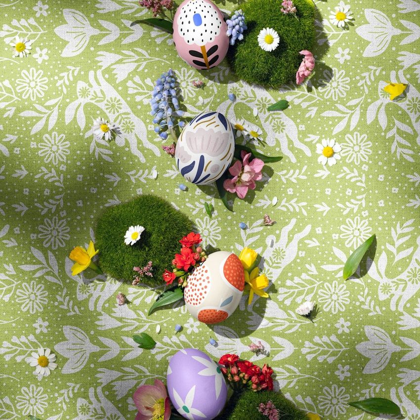 Easter eggs with colorful patterns are arranged on moss lumps and surrounded by spring flowers on a green patterned fabric.