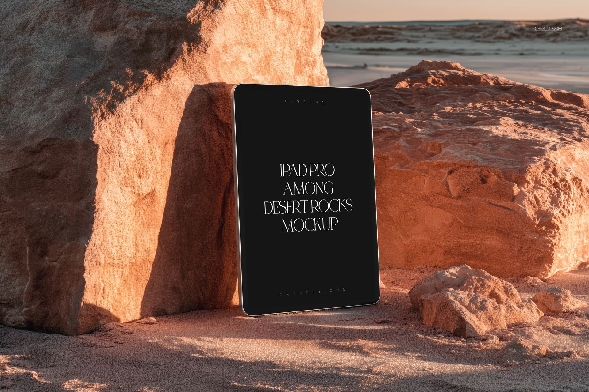 Front view of an iPad Pro mockup positioned among textured desert rocks, with sand and sunlight in the background.