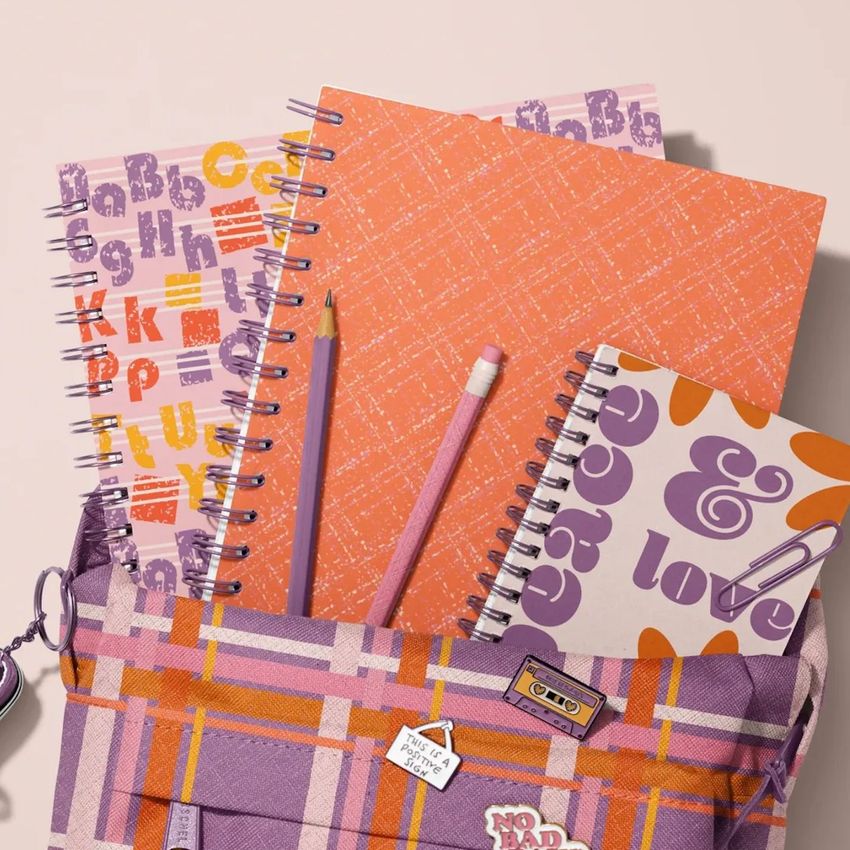 School backpack mockup with spiral notebooks, pencils, and patterned covers in orange, purple, and cream tones on a light surface.