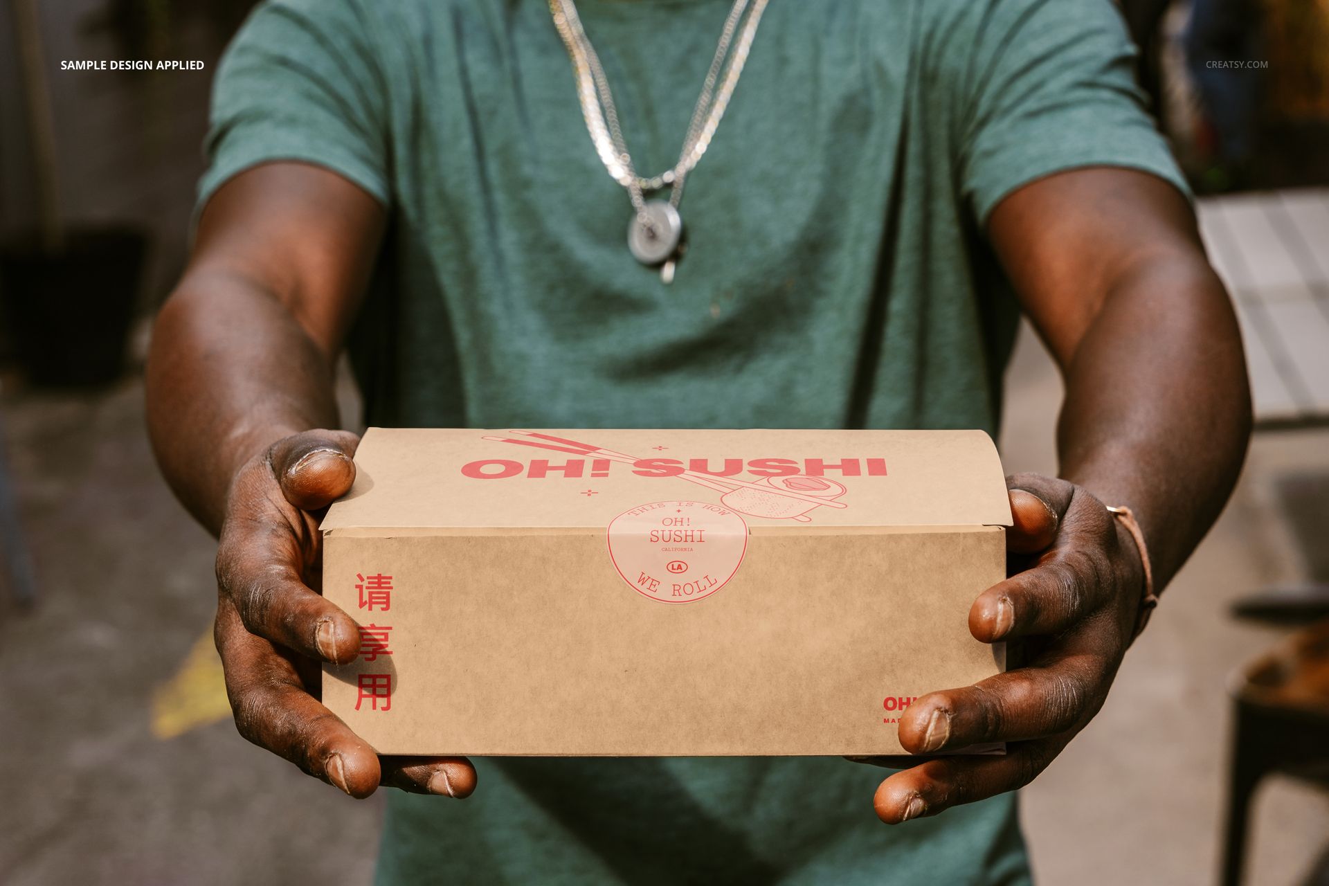 Close-up of hands presenting a kraft paper takeaway box mockup with red Asian-inspired design, outdoor environment.