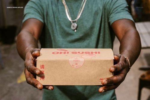 Close-up of hands presenting a kraft paper takeaway box mockup with red Asian-inspired design, outdoor environment.