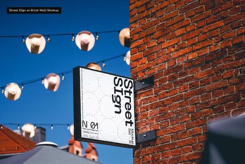 Rectangular street sign mockup mounted on a red brick wall, with string lights and blue sky in the background.