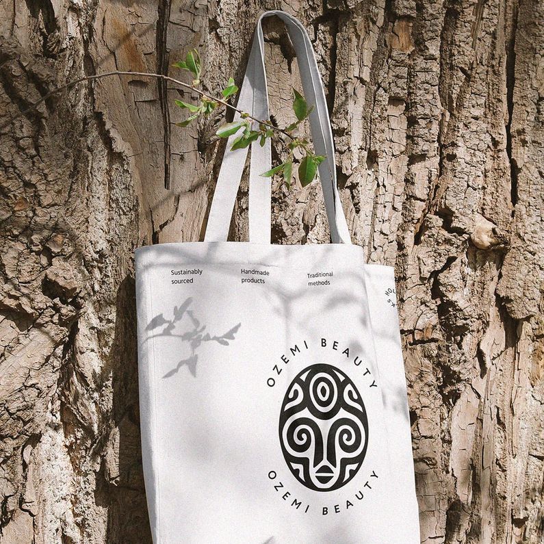 Tote bag mockup hanging on a textured tree trunk, featuring a bold black circular logo and text on a light fabric background.