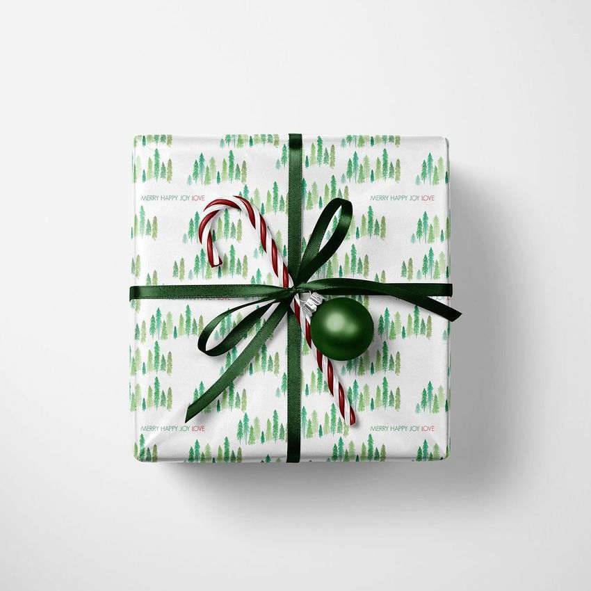 Christmas gift mockup featuring a square box wrapped in white paper with green tree patterns, tied with a black ribbon, a candy cane, and a green ornament on a white background.