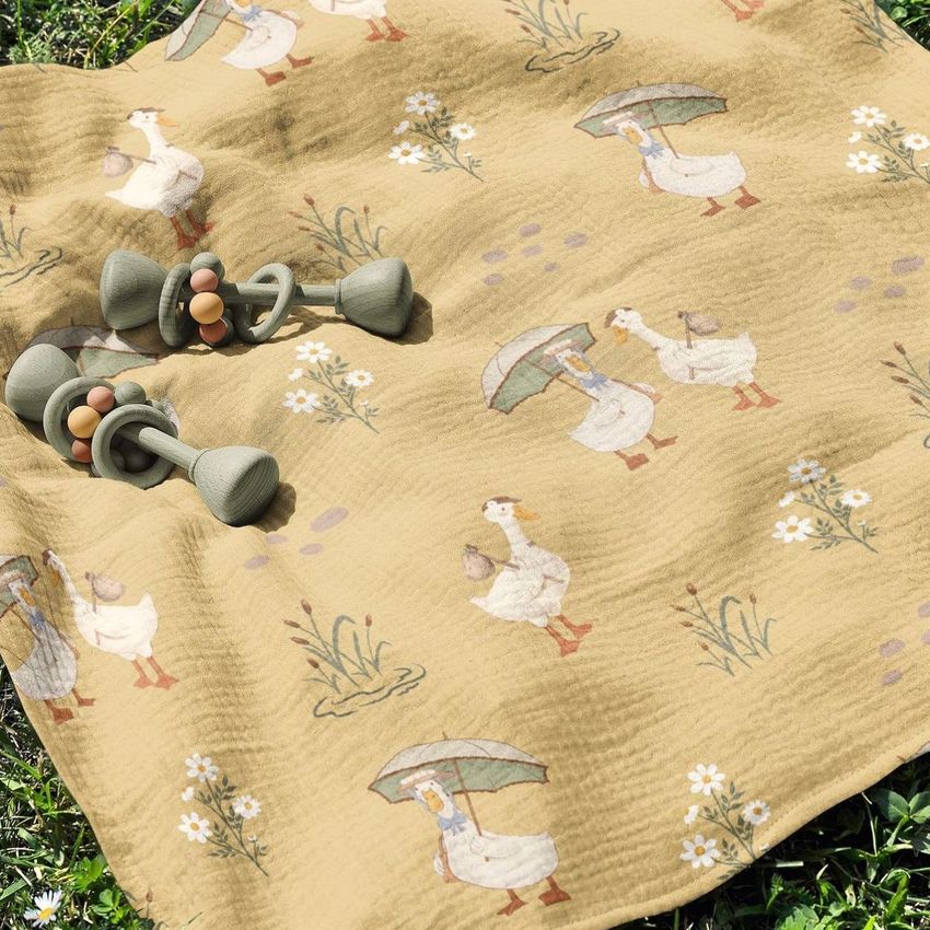 Crinkle cotton muslin swaddle blanket mockup featuring a yellow blanket with duck and floral illustrations, shown outdoors on grass with two green baby rattles.