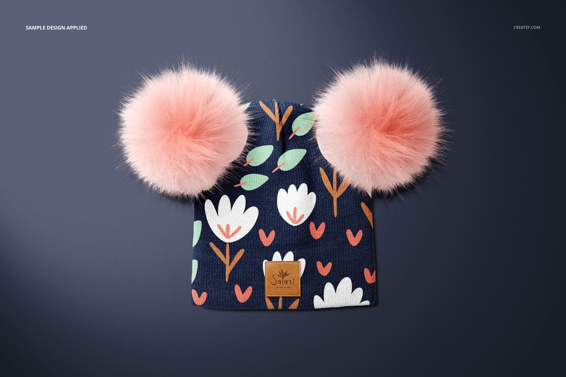 Children’s beanie mockup with fluffy pink pompons and white flower print, displayed on a smooth dark surface.
