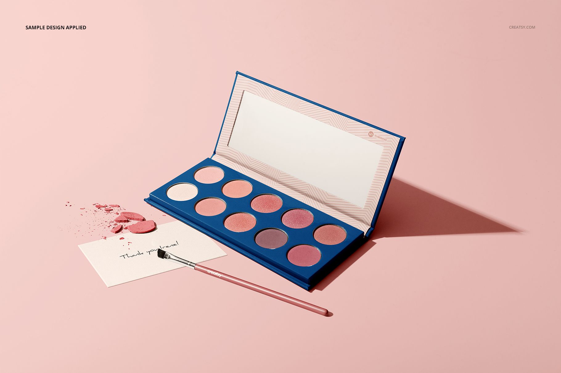 Open eyeshadow palette mockup featuring a blue case, pink-toned powders, makeup brush, and scattered pigment.