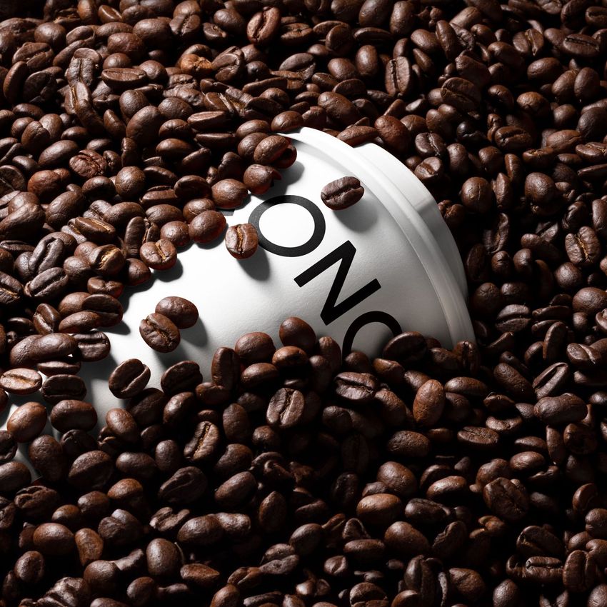 Paper cup mockup partially buried in a pile of dark roasted coffee beans, with bold black text visible on the cup.