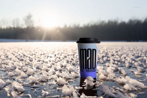 Winter-themed PSD mockup of a paper cup with a black lid placed on icy ground, sunlight in the background.