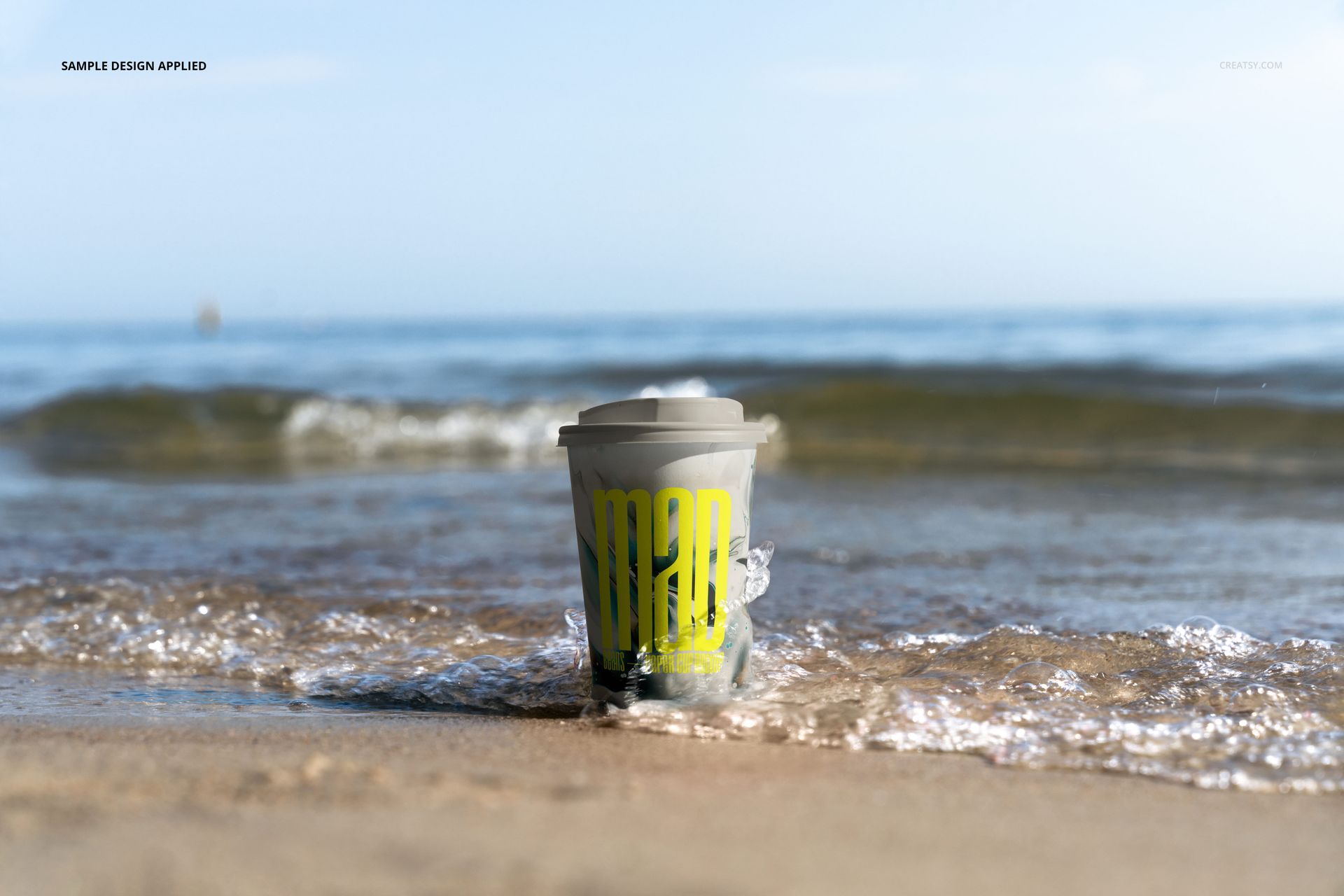 PSD mockup of a paper coffee cup with a gray lid and bright yellow print placed on sandy beach near the shoreline.