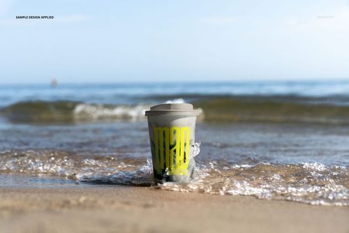 PSD mockup of a paper coffee cup with a gray lid and bright yellow print placed on sandy beach near the shoreline.
