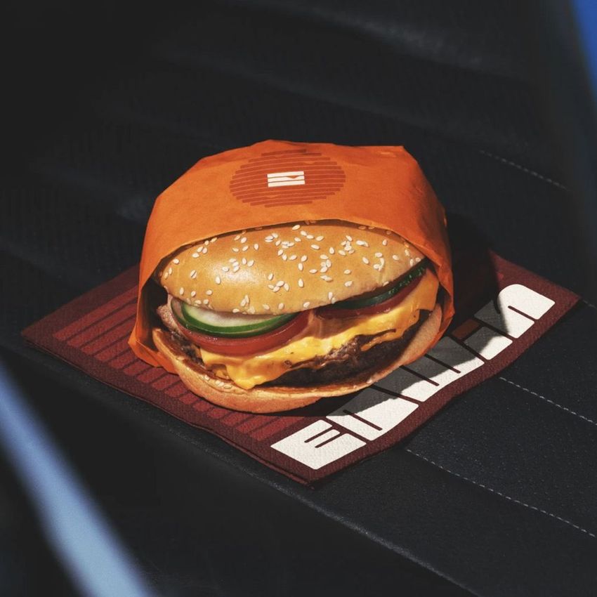 Burger mockup placed on a car seat, wrapped in orange paper with visible lettuce, tomato, and cheese, on a branded mat.