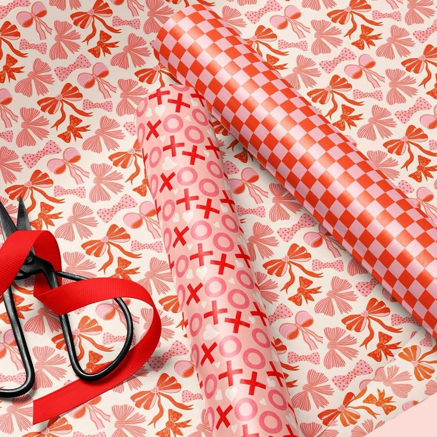 Gift wrapping paper mockup showing three rolls with red and pink patterns, scissors, and ribbon on a festive background.