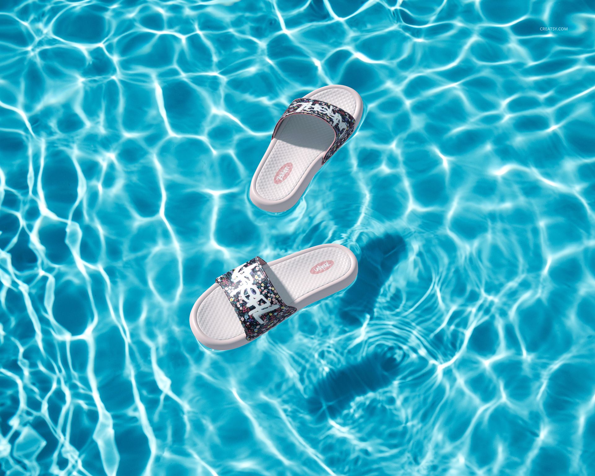 Two slide sandals with patterned straps floating on the surface of a sunlit swimming pool, top view mockup.