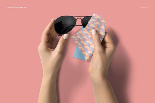PSD mockup showing hands cleaning black sunglasses with a patterned microfiber cloth against a pink surface.
