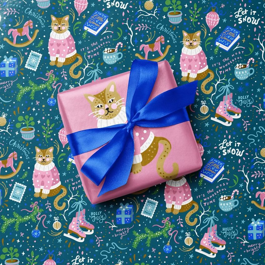 Square gift box mockup wrapped in pink paper with a cat illustration and blue ribbon, placed on festive patterned wrapping paper.