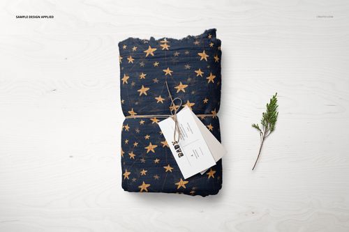PSD mockup of a muslin swaddle blanket in navy blue with yellow stars, displayed folded on a pale wooden background.