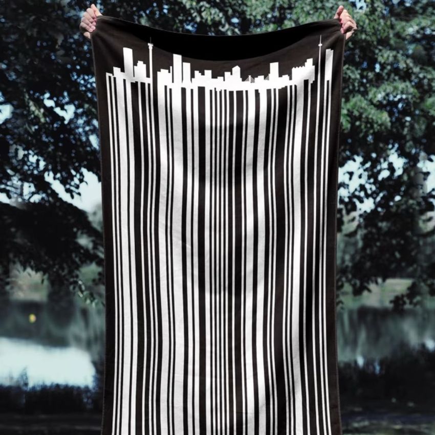 Beach towel mockup featuring a black towel with a white barcode design and city skyline, held outdoors in front of trees.
