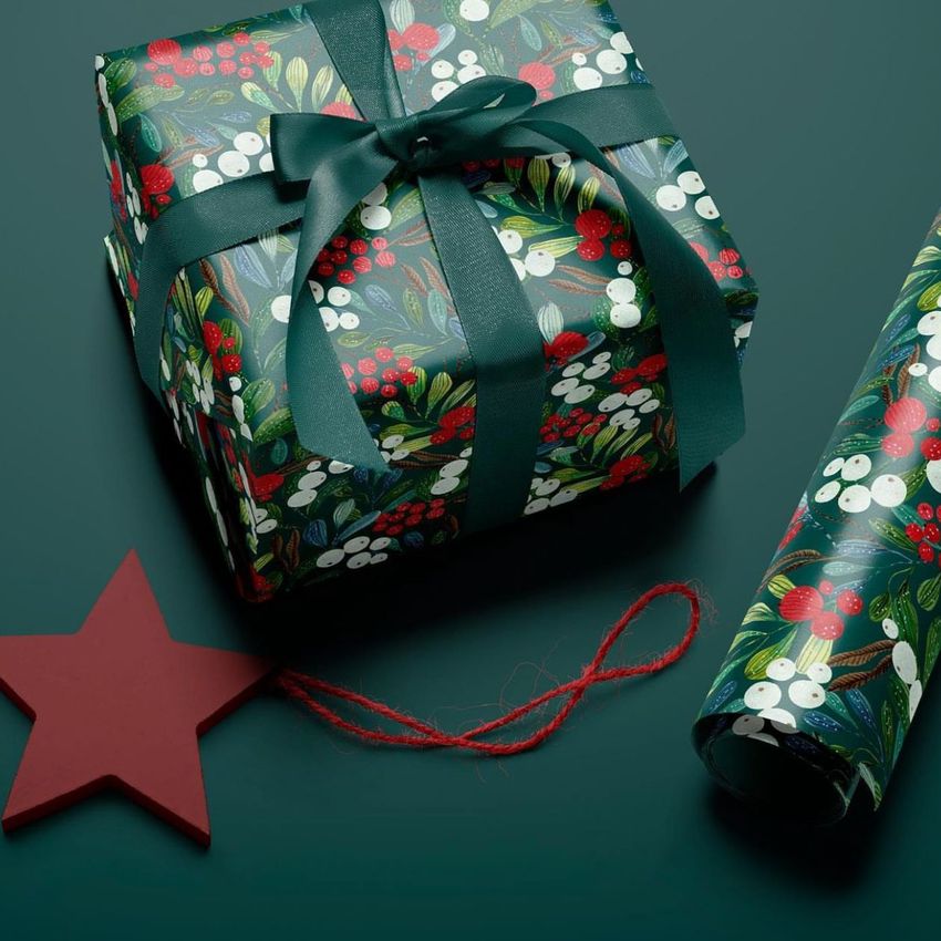Gift wrapping paper mockup featuring a wrapped box with a dark green ribbon, winter berries pattern, and a matching roll on a green surface.