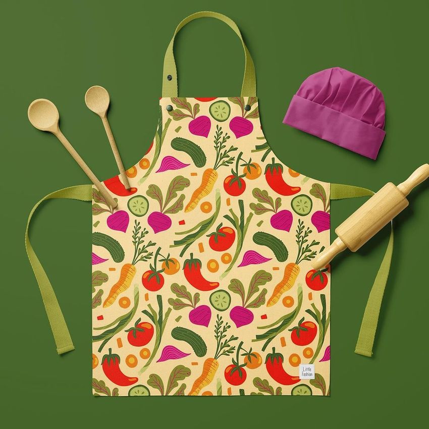 Kids apron mockup featuring a colorful vegetable pattern on a yellow background, displayed with a pink chef hat, wooden spoons, and rolling pin on a green surface.