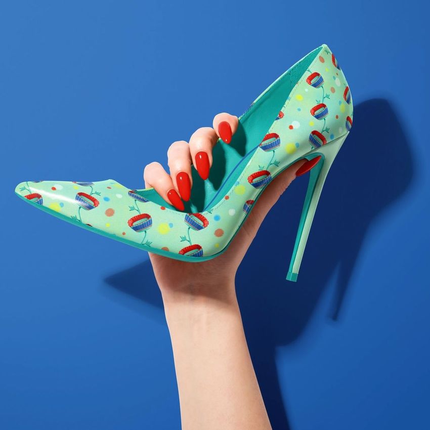 High heels mockup featuring a hand with red nails holding a turquoise stiletto decorated with colorful floral patterns against a blue background.