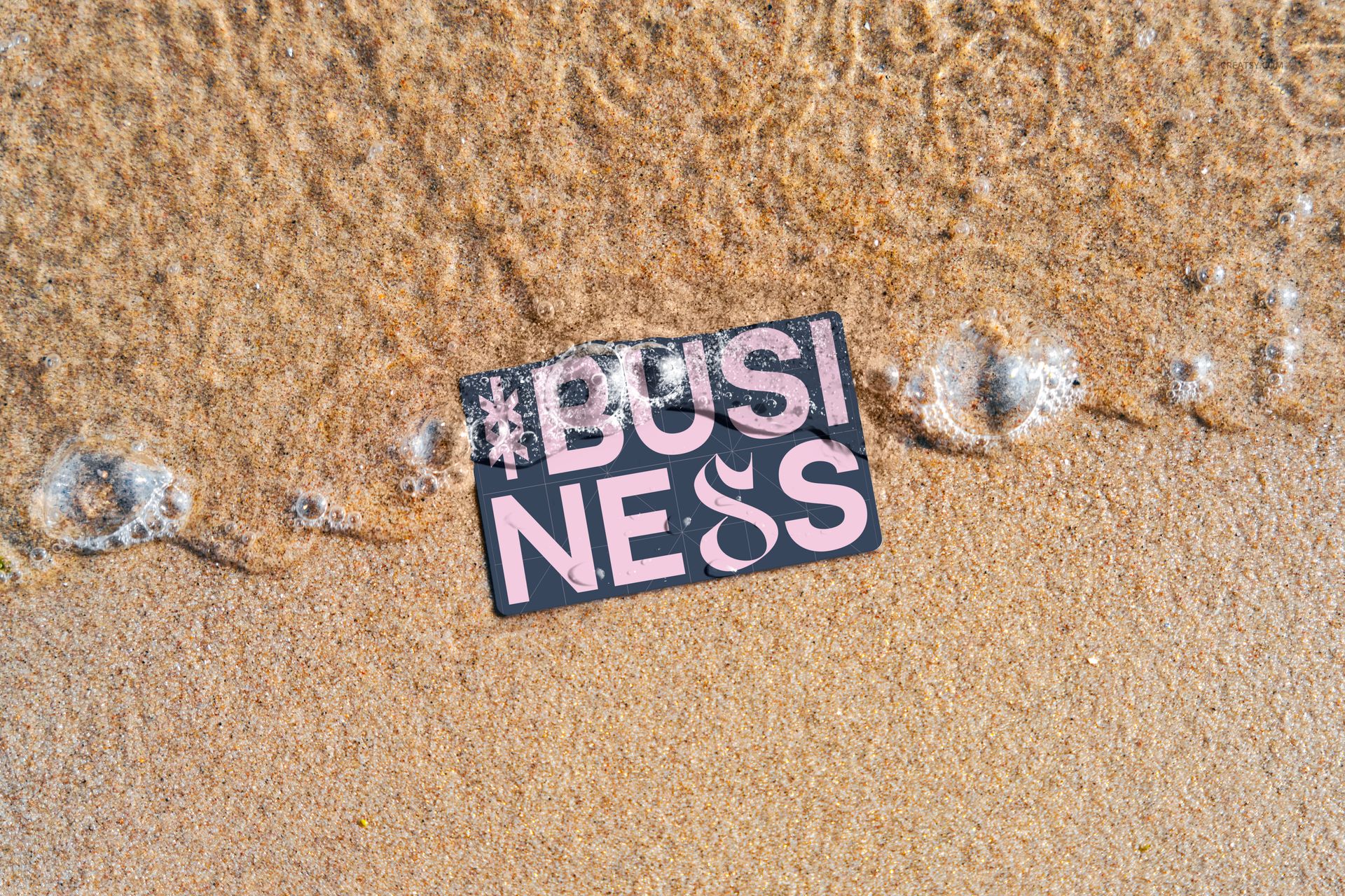Business card mockup placed on sandy beach with foamy water approaching, featuring large pink letters on a dark background.
