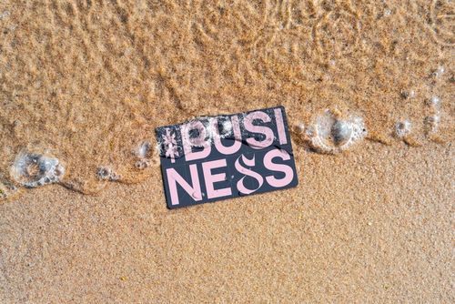 Business card mockup placed on sandy beach with foamy water approaching, featuring large pink letters on a dark background.