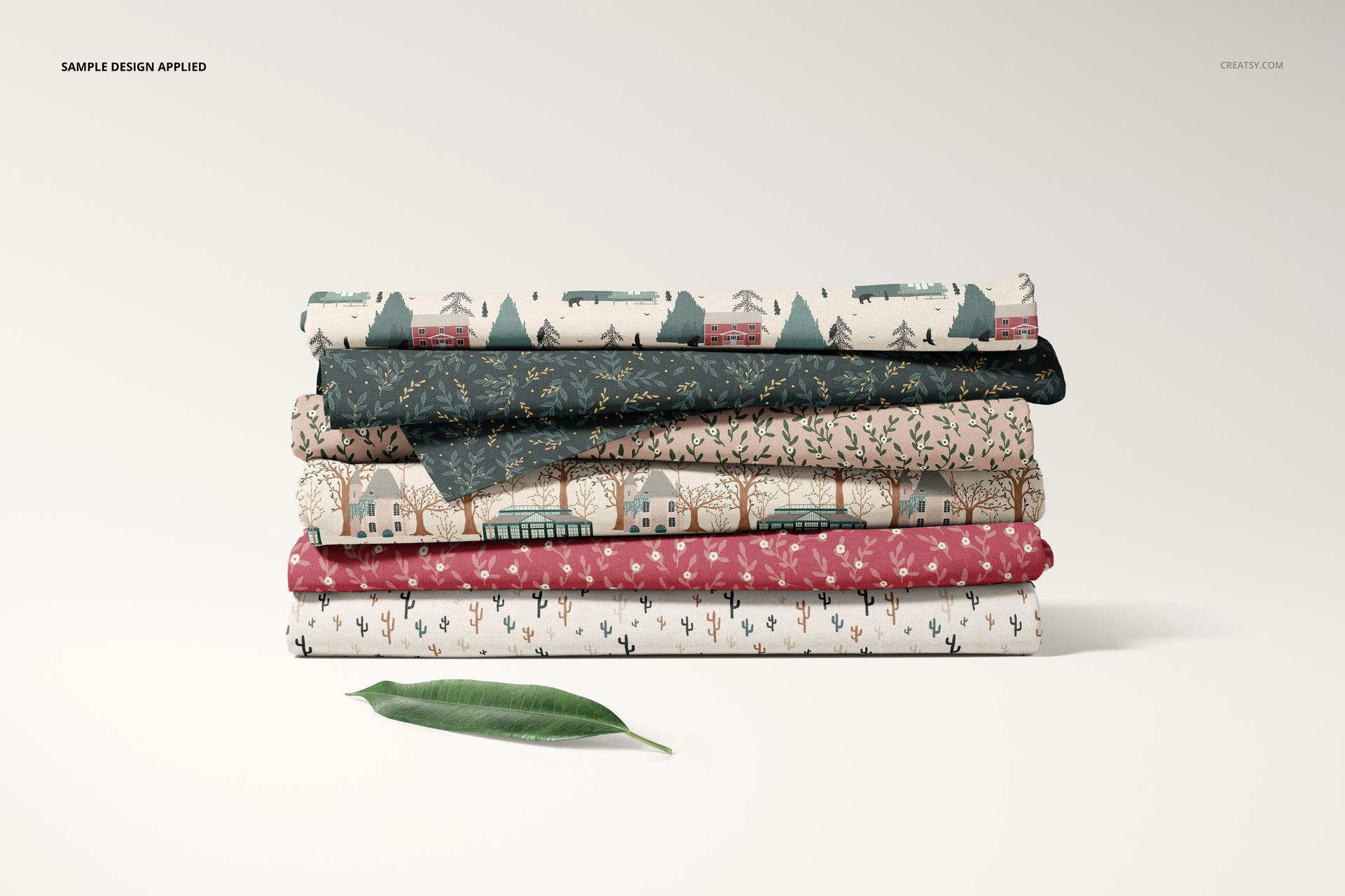 Front view of a fabric stack mockup showing assorted folded fabrics with botanical and geometric designs, leaf accent included.