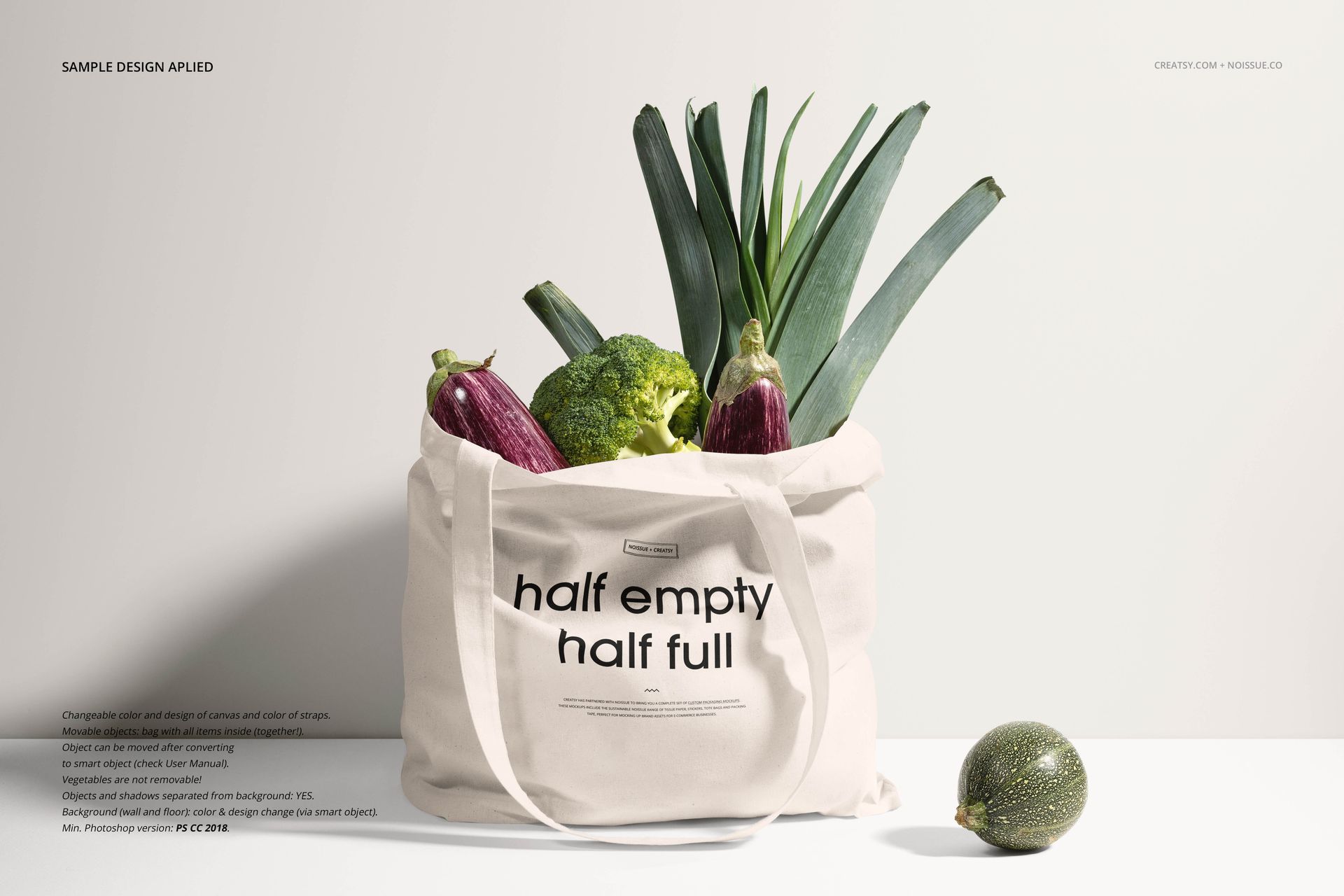 Front view of a canvas tote bag mockup filled with fresh vegetables, including broccoli, eggplant, and leek, on a light surface.