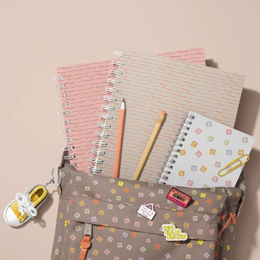 School backpack mockup with spiral notebooks, pencils, and a sneaker keychain arranged on a beige surface.