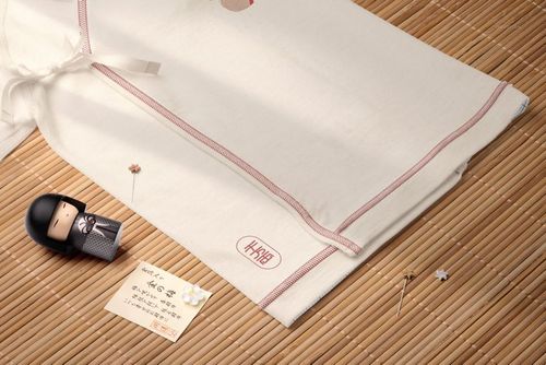 Organic cotton baby kimono bodysuit mockup displayed on a bamboo mat with a doll, note, and small pins.