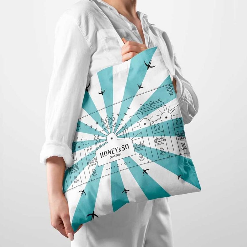 A person in a white shirt holds a tote bag mockup featuring a blue and white geometric design with stylized buildings and text.