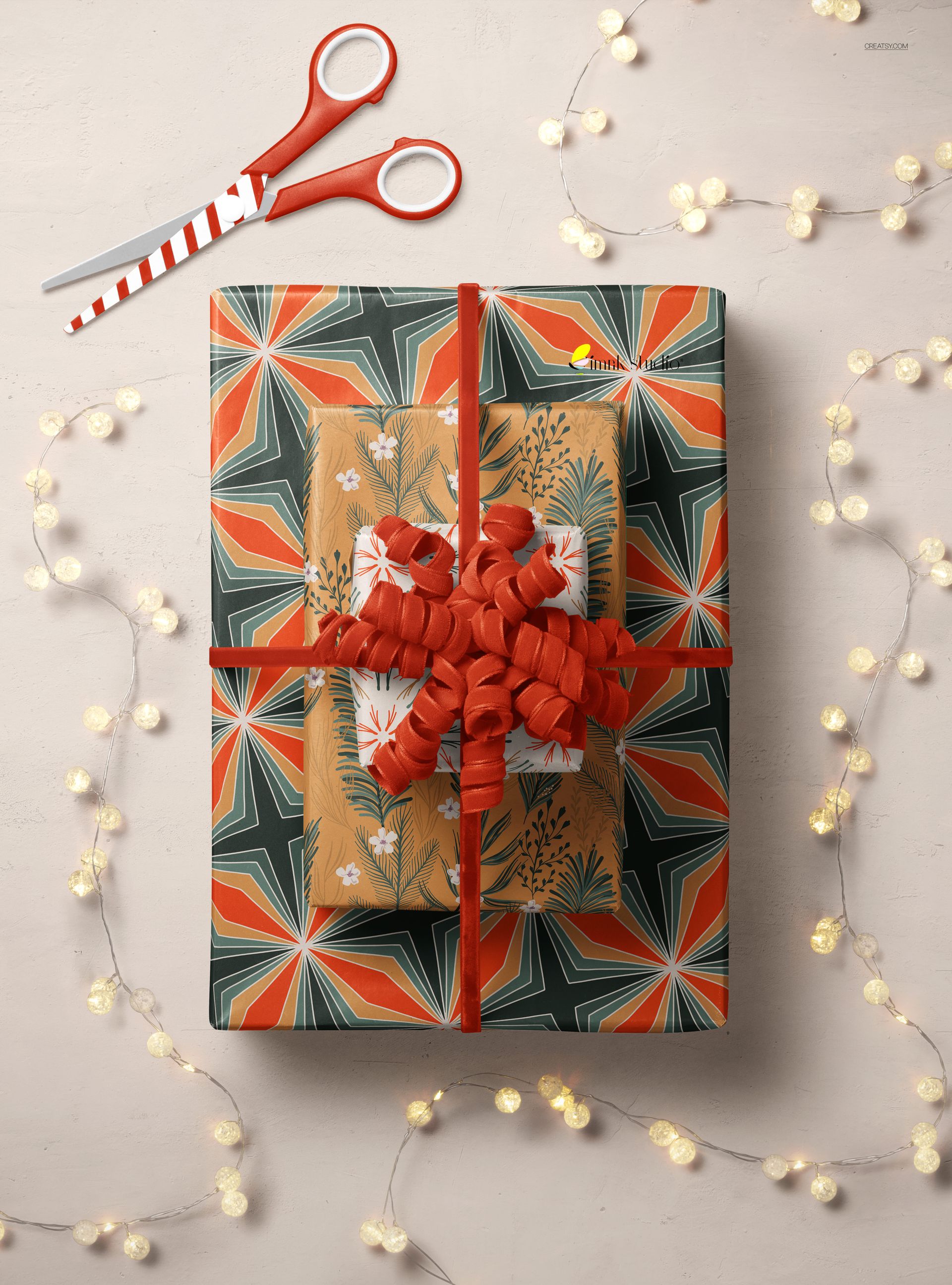 Christmas-themed wrapped gift box mockup with bold red ribbon, surrounded by string lights and scissors, top view.