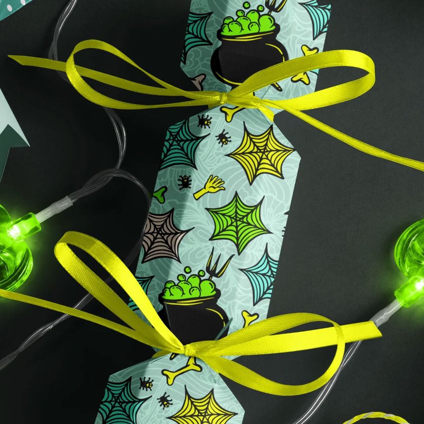 Halloween party cracker packaging mockup with a light blue wrapper featuring cauldrons, spider webs, and bones, tied with yellow ribbons.