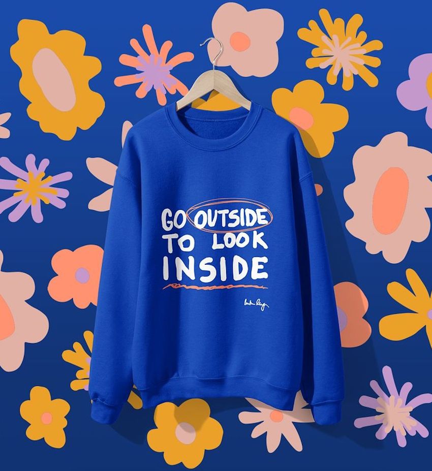 Blue sweatshirt mockup hanging on a wooden hanger against a blue wall with colorful naive art floral patterns.