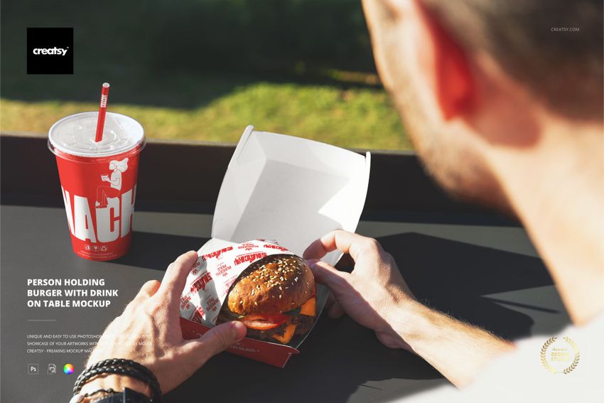 Front perspective of hands holding a wrapped burger next to a red soda cup, with packaging mockup elements on a table.