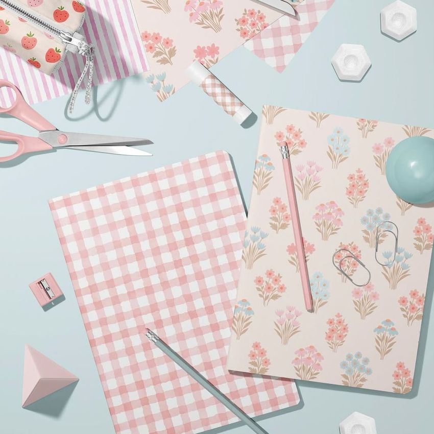 Stationery mockup featuring two classic A4 notebooks, one with a pink gingham cover and one with a pastel floral pattern, surrounded by scissors, pens, and paper clips on a light blue surface.