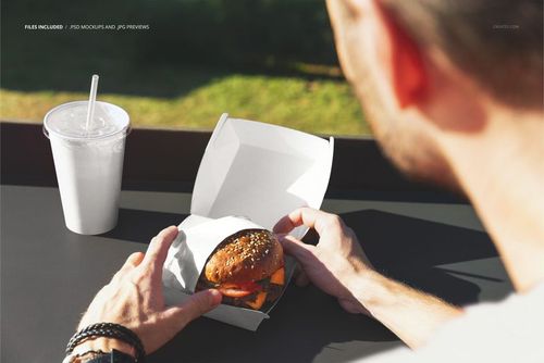 Close-up of hands opening a burger box next to a plastic cup with straw on a sunlit outdoor table, PSD mockup scene.
