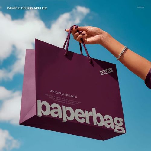 PSD mockup of a purple paper shopping bag with large lettering, held up against a sky with scattered clouds.